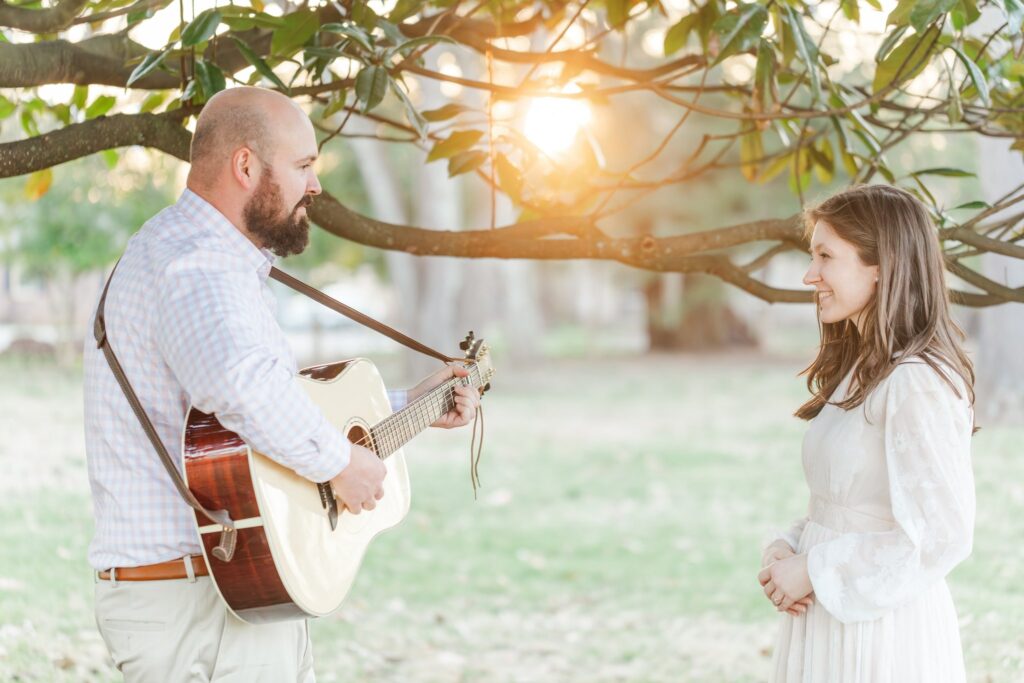 man playing guitar under magnolia tree with a lady in a white dress watching him