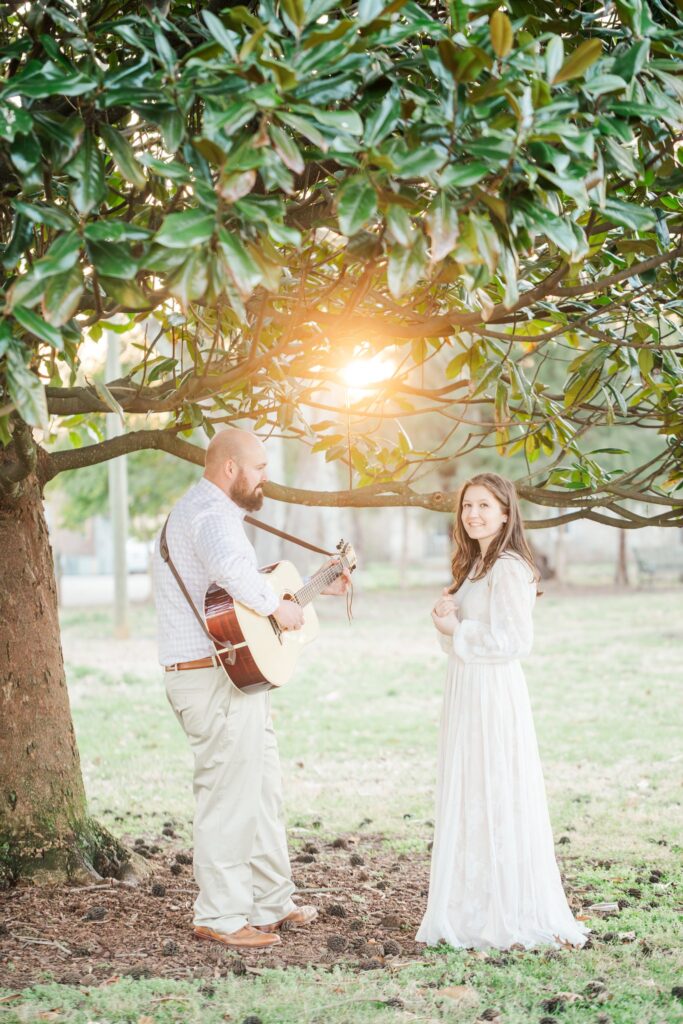 man playing guitar under magnolia tree with a lady in a white dress watching him