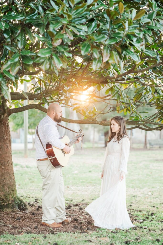 man playing guitar under magnolia tree with a lady in a white dress watching him