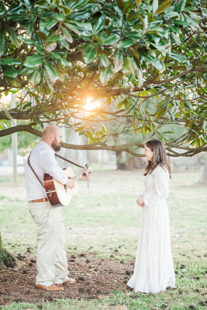 man playing guitar under magnolia tree with a lady in a white dress watching him
