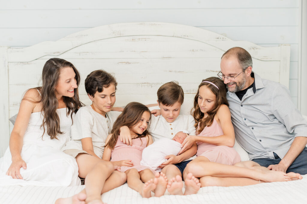 Family of seven sitting on their white bed smiling at their newborn baby! 